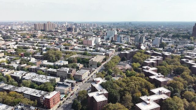 A broad aerial sweep moves over Brooklyn residential blocks, apartment buildings and tree-lined streets in daylight. Green courtyards and outdoor courts break up the dense pattern of city streets.