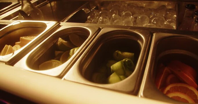 Close up of fresh lemon and lime wedges arranged in stainless steel garnish trays beside ice cubes. Warm golden lighting illuminates the cocktail preparation station at a bar.