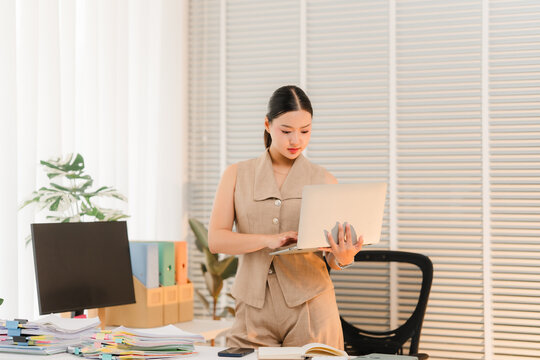 Office Professional Typing on Laptop: A focused businesswoman stands in a modern office, engrossed in her work on a laptop, surrounded by organized desk essentials and natural light.