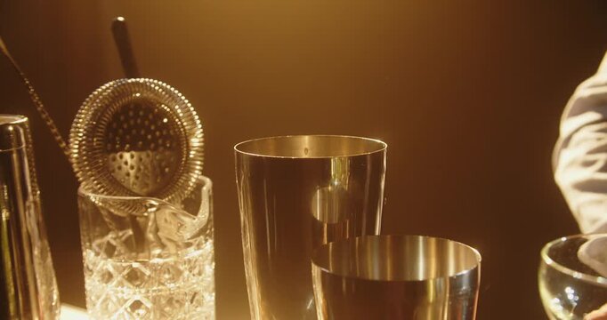 Close up of a bartender dropping a large spherical ice ball into a stainless steel cocktail shaker. Warm golden lighting highlights the bar tools and drink preparation process.