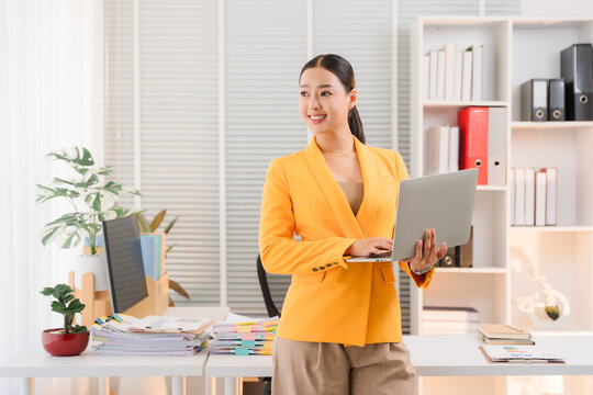 Businesswoman with Laptop in Office: A smiling businesswoman stands confidently in a modern office, holding a laptop and looking towards the side with a determined yet pleasant expression.