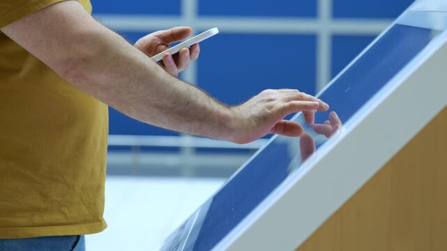 Man interacts with touch screen kiosk. He holds phone in one hand. Background shows blue paneled wall. Scene captured indoors with bright lighting. Focus on hands and screen interaction