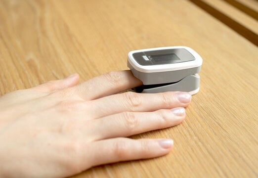 Blood oxygen saturation reading from pulse oximeter clipped on finger. Hand rests on wooden table while fingertip sensor tracks heart rate and vitals. Portable digital gadget for home health