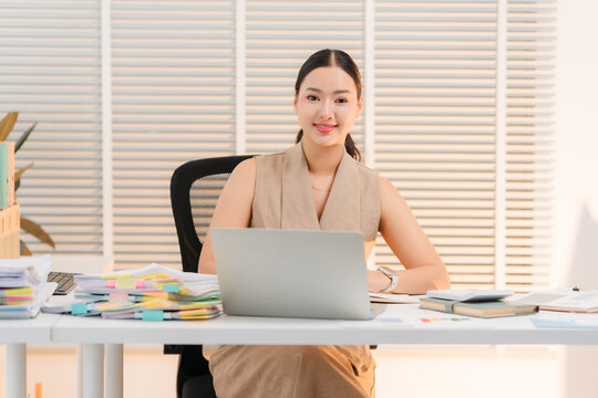Asian Woman Working at Desk: A poised Asian businesswoman sits at her desk, laptop open and documents neatly stacked, ready for a productive day in her well-lit office.
