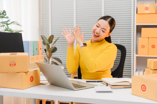 Excited Entrepreneur: A young woman enthusiastically claps her hands while working in her home office, surrounded by packages ready for shipping.