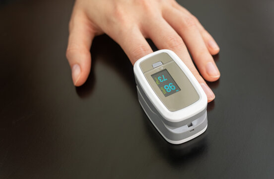 White pulse oximeter clipped on finger, oxygen saturation 98 and pulse rate 73. Fingertip sensor monitors SpO2 and heart rate, hand resting on dark glossy table. Top view, gadget for self