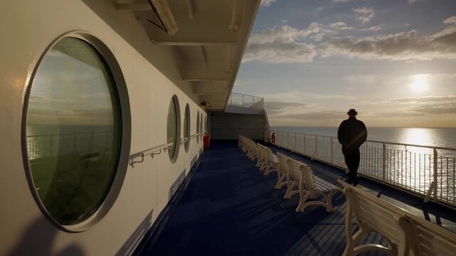 Person stands by a ferry railing in low sunlight beside rows of benches and round cabin windows