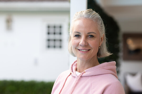 Middle-aged woman standing on porch by paned window wearing pale pink hoodie and stud earring