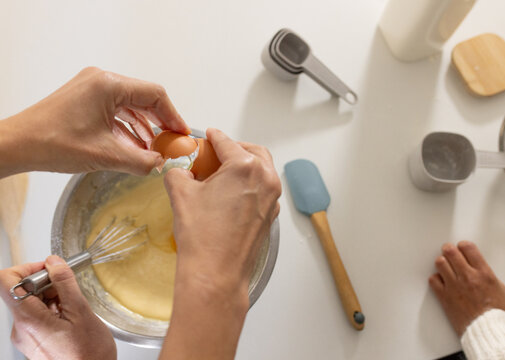 Female friends cracking egg and whisking batter on white countertop with metal mixing bowl
