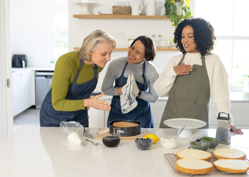 Diverse female friends gathering at kitchen island wearing aprons inspecting springform cake