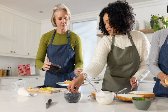 Diverse female friends wearing aprons decorating cakes with frosting at island in bright kitchen
