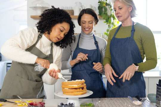 Diverse female friends wearing aprons, piping with bag, assembling layer cake on rotating stand