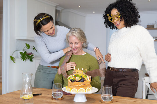 Female friends with senior woman wearing sweaters, placing sash with fruit cake in kitchen