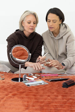 Two women wearing hoodies sitting on rust-orange blanket at home, sharing compact and brush