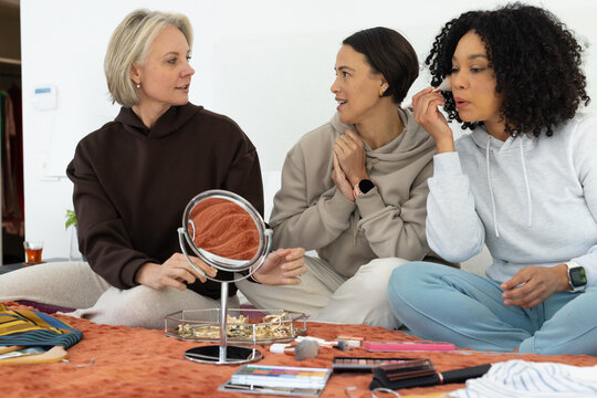 Diverse female friends sitting on bed in loungewear applying mascara using tabletop mirror