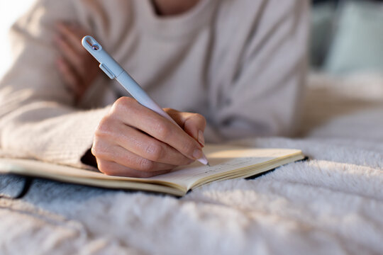 Female writing in small notebook on plush bedspread, holding light-gray pen, wearing knit sweater