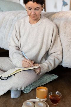 Woman 30s sitting on bedroom floor wearing knit loungewear writing in open notebook with tea