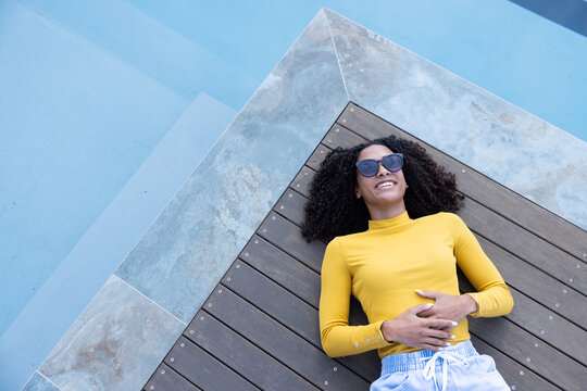 African American woman lying on deck with screws by pool, wearing mustard top, shades, smiling