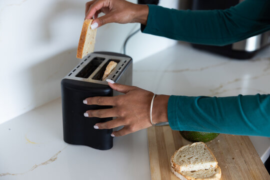 Hands placing slice of bread into black two-slice toaster on marble countertop near cutting board
