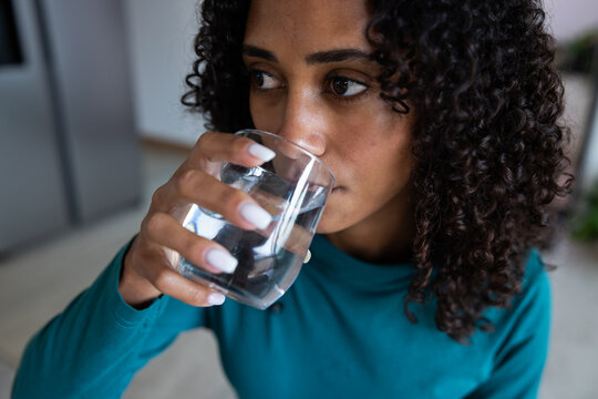 African American woman holding clear glass, sipping water in kitchen wearing teal long sleeve top