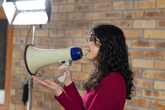 Woman in maroon top holding megaphone, facing right, gesturing in studio by softbox and brick wall