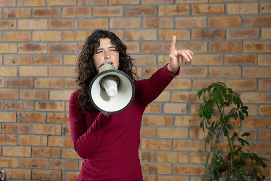 Adult female in burgundy top by exposed brick wall, holding megaphone, pointing, plant to right