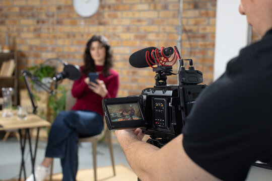 Presenter and operator reading on chair with smartphone in studio while filming with cinema camera