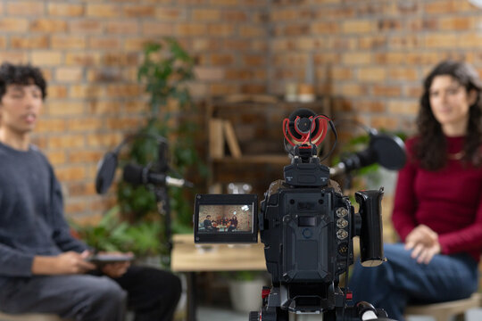 Two hosts sitting in small studio filming conversation with camera flip out LCD and microphones