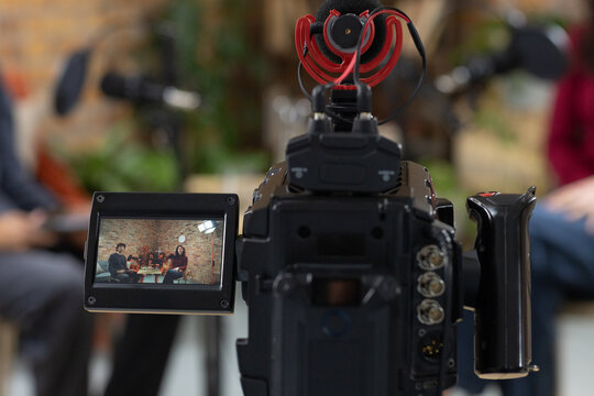 Camera dominating foreground, showing flip-out monitor with three on couch, red mic on talk set