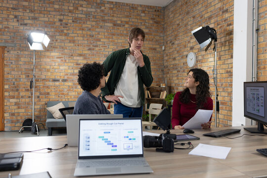 Diverse coworkers discussing storyboard at brick-walled studio table with laptop printed pages