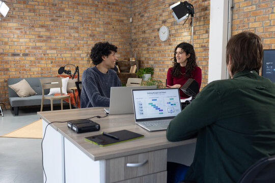 Diverse coworkers woman in burgundy top discussing editing in studio with laptops and tablet