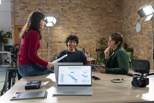 Diverse coworkers collaborating at table in loft studio, reviewing editing laptop, DSLR, tablet
