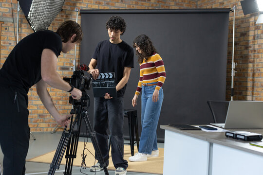Production crew adjusting camera, holding clapperboard, talent wearing striped sweater at studio