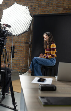 Adult woman sitting on black stool in studio, facing camera with softbox, laptop, cables