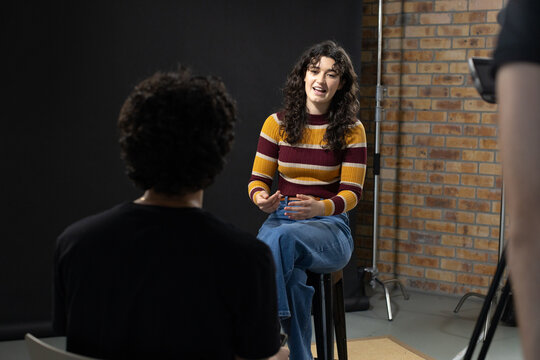 Female interviewee on tall stool gesturing and speaking to listener and crew in studio striped knit