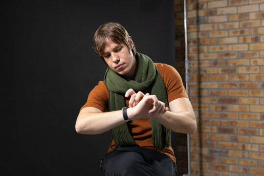 Man wearing rust knit top and olive scarf adjusting black wristwatch sitting on stool in studio