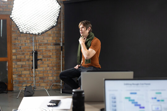 Adult male 20s sitting on stool in studio wearing rust top and olive scarf under softbox