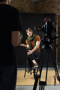 Diverse crew filming man in orange top green scarf sitting in brick studio with camera tripod