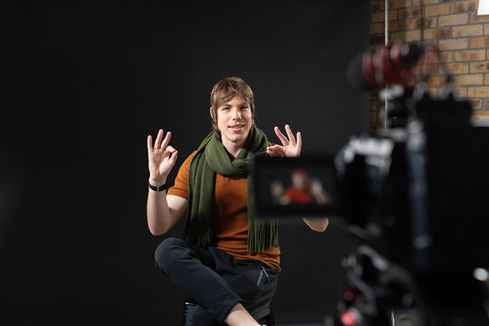 Male sitting on stool in studio beside camera monitor, wearing orange shirt, making OK gesture
