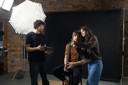 Studio creative team adjusting model seated on stool wearing rust top on set with softbox, tablet