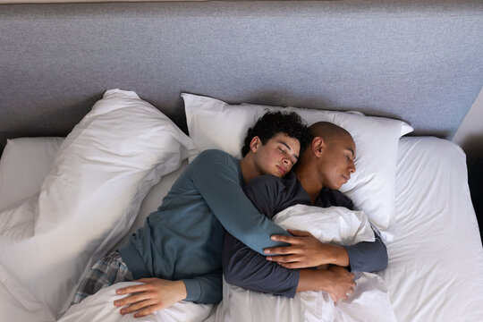 Two men cuddling in bedroom on bed with white duvet, pillows, grey headboard, wearing pajamas