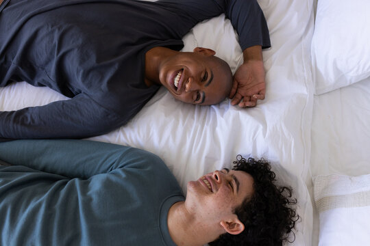 Male friends lying in bedroom on bed with white pillows and duvet, African American man smiling
