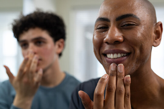 Male friends, bald man touching chin in bathroom while roommate touching upper lip by blinds