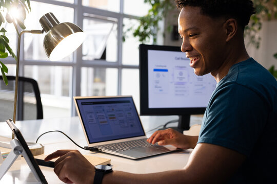 African American man sitting desk by window, wearing teal shirt, using laptop, tablet, copy space