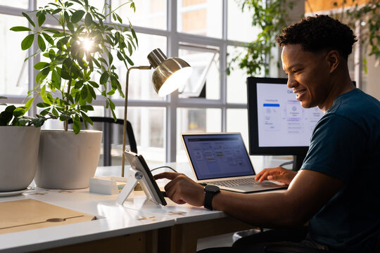 African American man wearing teal tee sitting using laptop and tablet at office, copy space