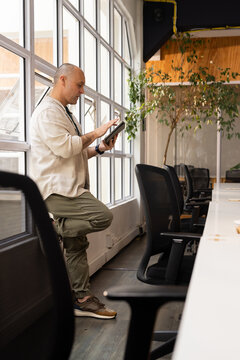 Adult man leaning against windowsill and using tablet at coworking space by grid windows