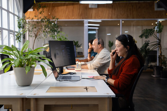 Diverse coworkers speaking into headsets at shared desk using laptop and large monitor, copy space