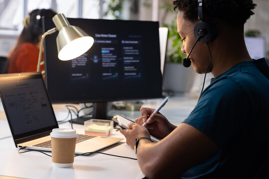 Diverse coworkers collaborating at open plan office with headset, laptop, monitor, coffee cup