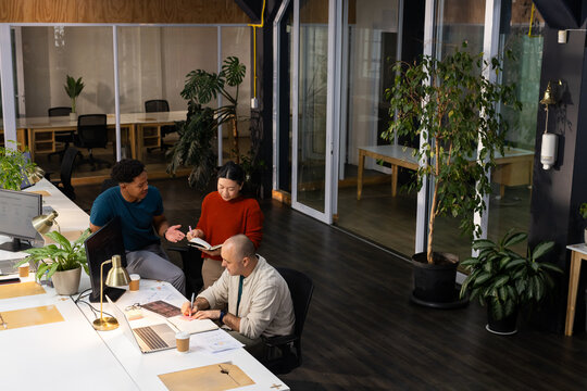 Diverse team in business attire gesturing writing leaning at desk in office with laptop notebook