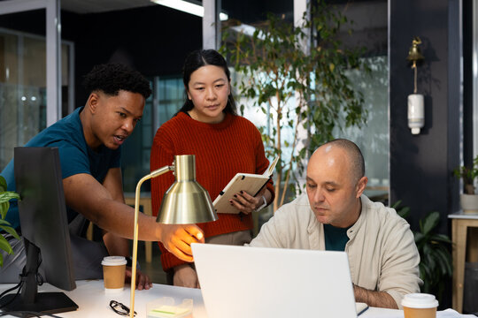 Diverse coworkers leaning over laptop at modern office workstation, pointing at brass desk lamp